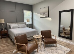 Een slaapkamer met een bed, twee stoelen, een ronde tafel, een lamp, een spiegel en een schilderij aan de muur.