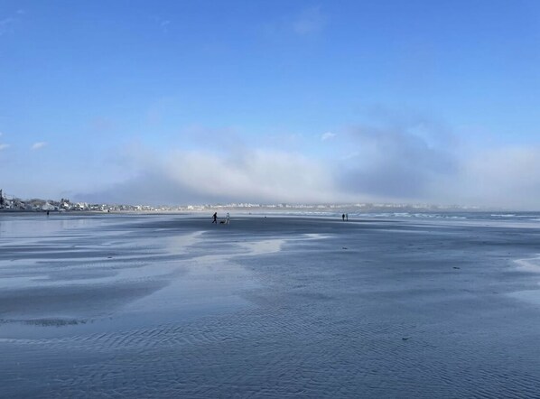 Une vaste plage de sable plat, sous un ciel dégagé, avec des bâtiments visibles au loin.