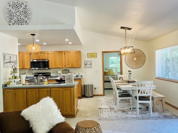 A modern kitchen with wooden cabinets, a central island, and a dining area with white chairs and a table. - Riverfront 3 bed/2 bath home, relaxing and peaceful, new furnishings and paint (Gasquet)