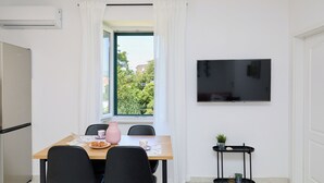 A modern kitchen with a built-in oven, a dining table with chairs, a TV mounted on the wall, and a window with curtains.