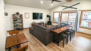 Ein gemütlicher Wohnbereich mit einer Couch, einem Holztisch und Barhocker. Ein Fernseher ist an der Wand befestigt, und ein Deckenventilator ist sichtbar.