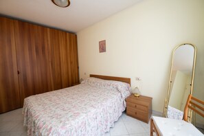 Quarto com guarda-roupa de madeira, cama com colcha florida, mesa de cabeceira com abajur e espelho de corpo inteiro.
