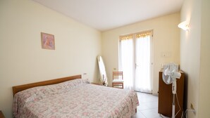Ein Schlafzimmer mit einem Bett, einem Standventilator, einem Stuhl, einem Spiegel und einer Tür mit Vorhängen.