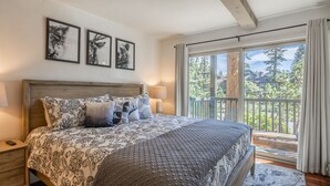 A bedroom with a large bed, a nightstand, a window with a view of trees, and three framed pictures on the wall.