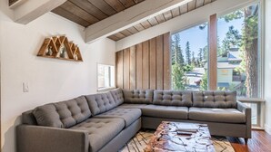 A modern living room with a large sectional sofa, wooden ceiling beams, and a large window offering a view of trees and buildings.