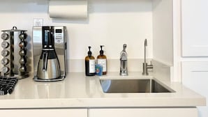 Una cocina con una cafetera de acero inoxidable, un hervidor, un lavabo y dos botellas de jabón para manos.