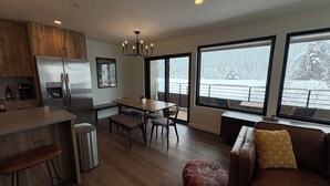 Una cocina moderna con un refrigerador de acero inoxidable, armarios de madera y un comedor con una mesa y sillas de madera. Hay un ventanal que ofrece vistas de una terraza exterior nevada.