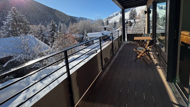 Un balcón con piso de madera, barandilla de metal y una mesa con sillas. Se observa un paisaje montañoso nevado con árboles y edificios.