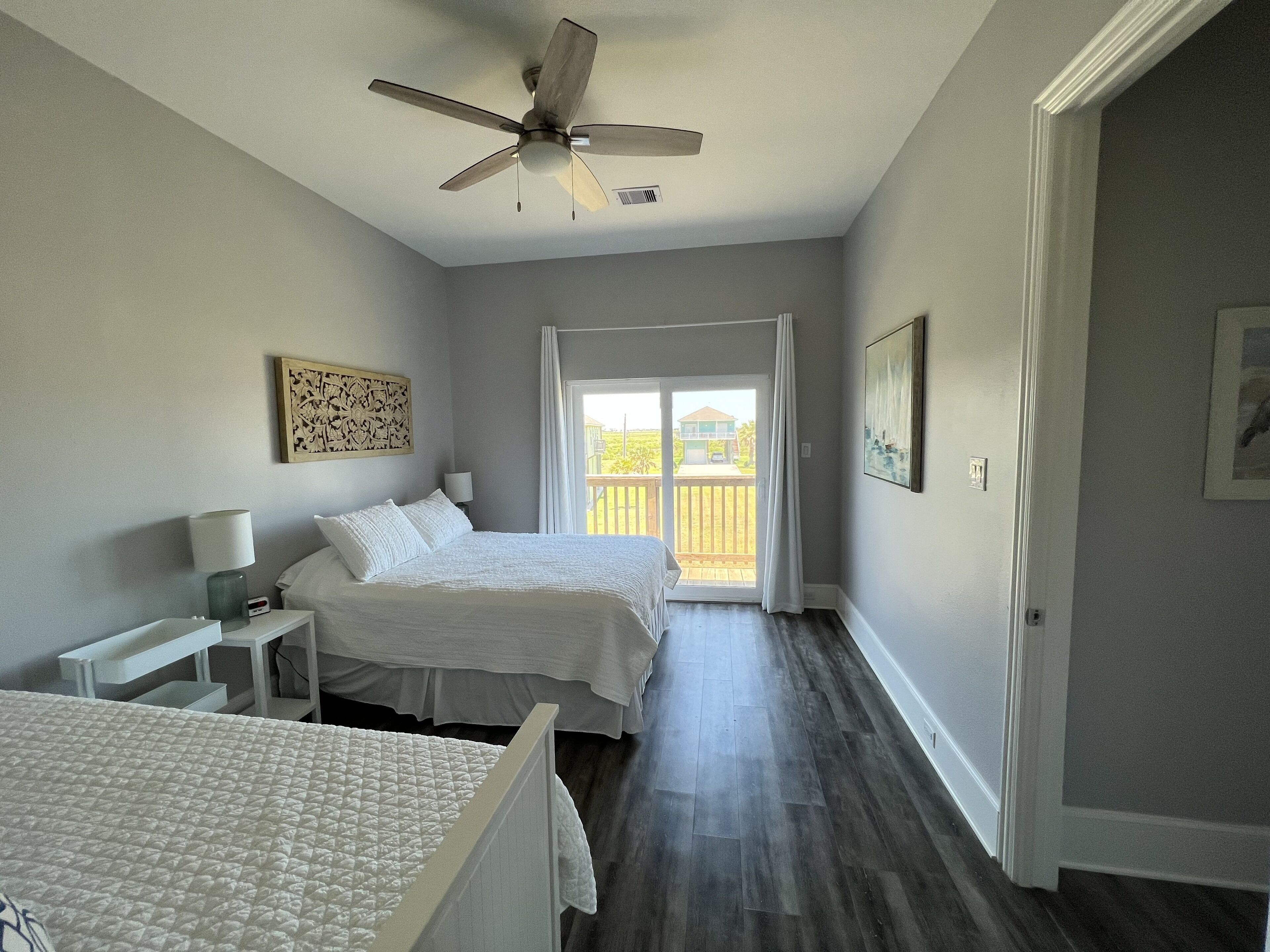 Quarto com cama, mesa de cabeceira, ventilador de teto e vista para o exterior por uma porta de correr.