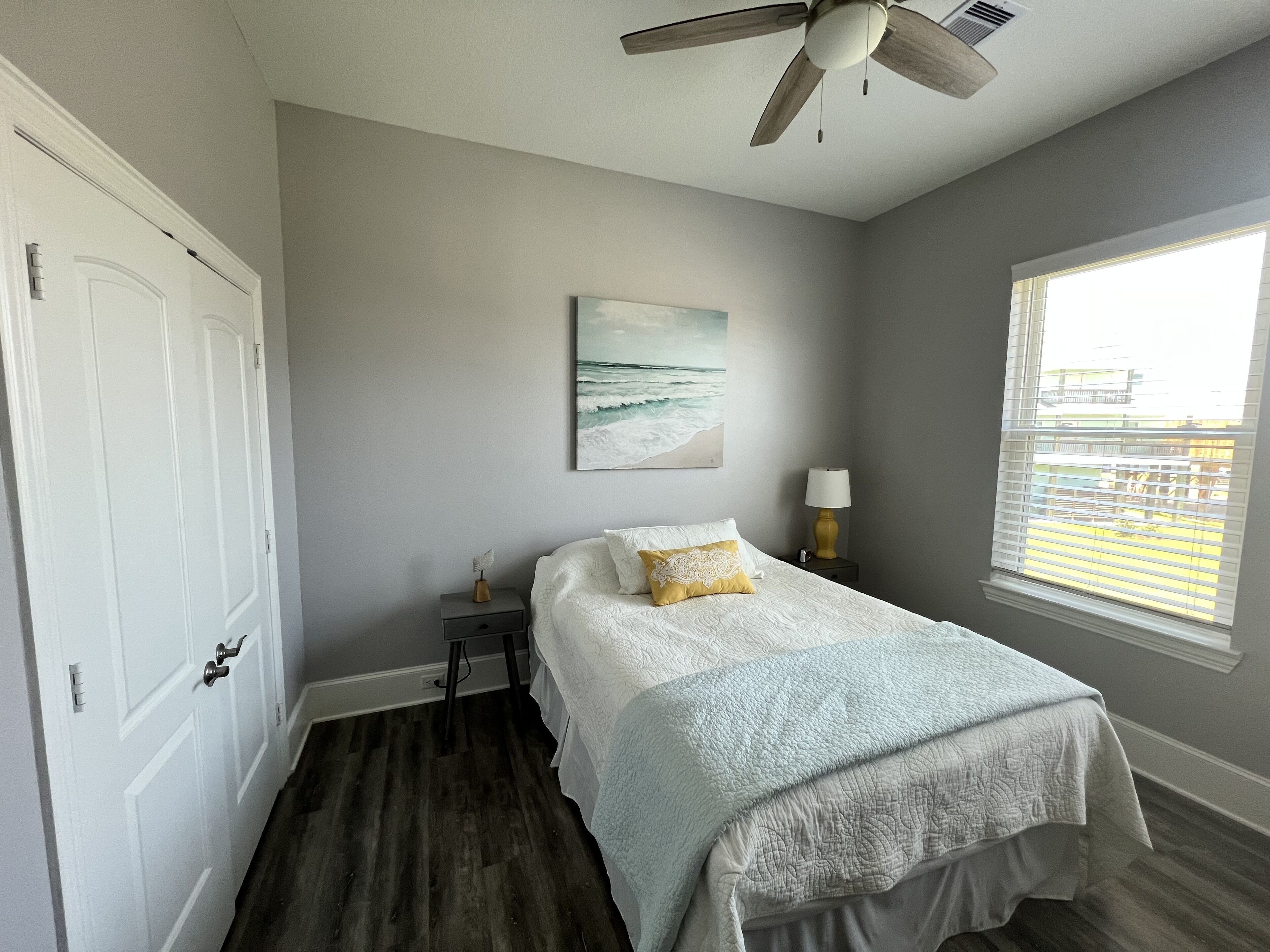 A bedroom with a bed, a ceiling fan, a painting, a nightstand, and a window.