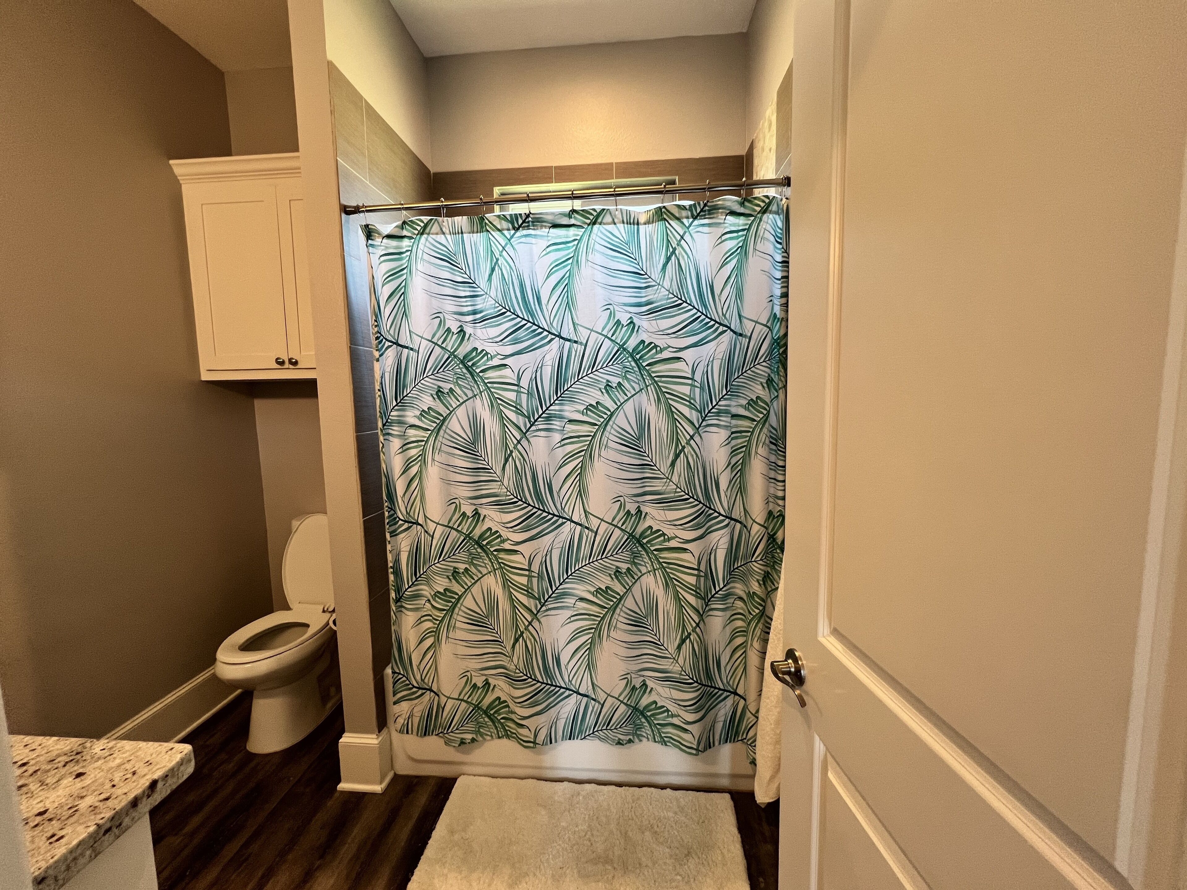 A bathroom with a shower curtain featuring a tropical leaf pattern, a toilet, and a cabinet above the toilet.