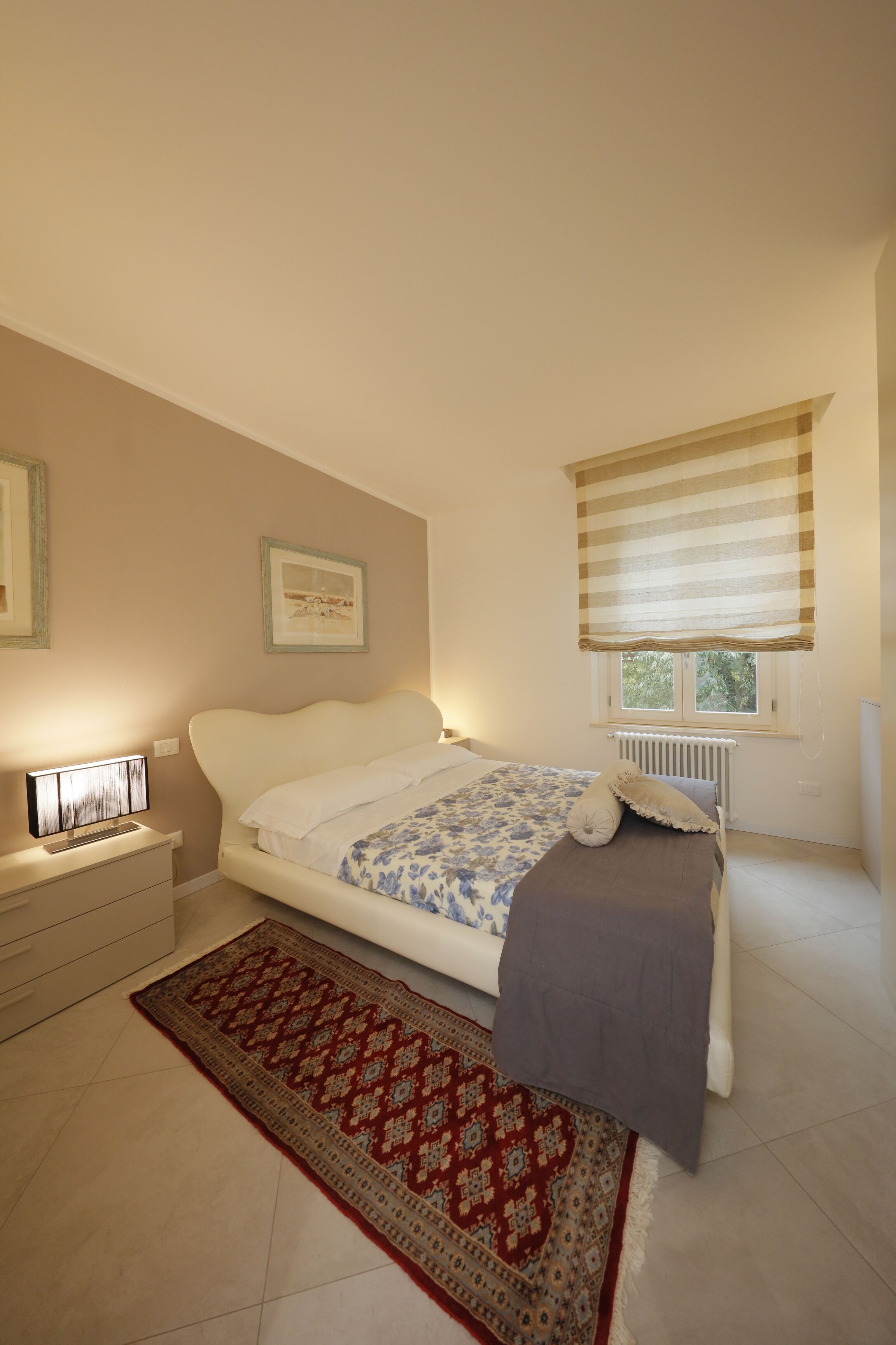 A bedroom with a bed, a nightstand, a window with blinds, and a radiator.
