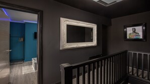 Ein Korridor mit einer dunklen Wand, einem weißen, verzierten Spiegel und einem gerahmten Bild mit Text.