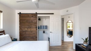 Une chambre avec un lit, une porte coulissante en bois, un ventilateur de plafond blanc, un couloir avec une porte, et une plante posée sur une console.