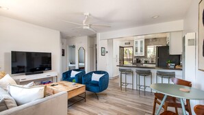 Un salon moderne avec un téléviseur à écran plat, un canapé, une table basse et deux fauteuils bleus. On y trouve également un coin repas avec une table et des chaises. La cuisine est visible au fond, avec des meubles blancs et un réfrigérateur noir.