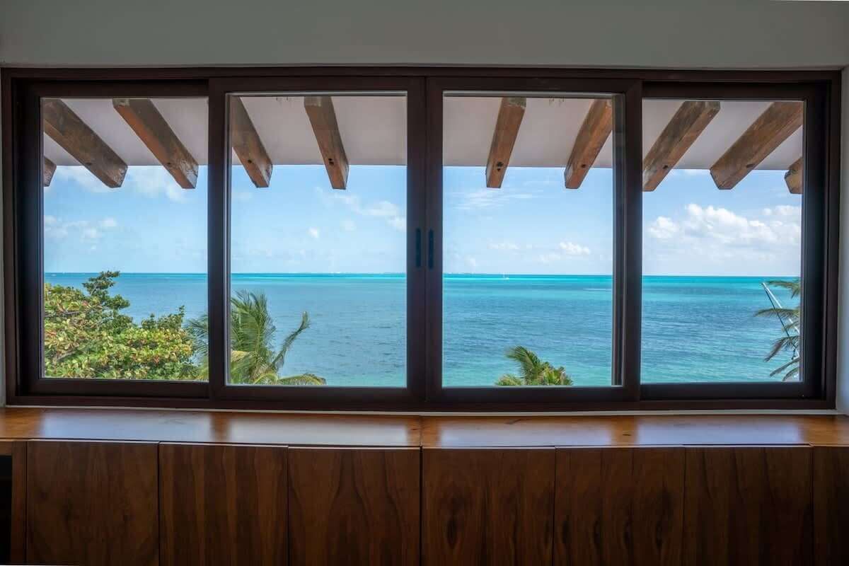 Een kamer met een groot raam met uitzicht op een turquoise zee en heldere lucht.