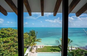 Une vue depuis une fenêtre donnant sur une mer bleue et limpide, une piscine et une végétation luxuriante.