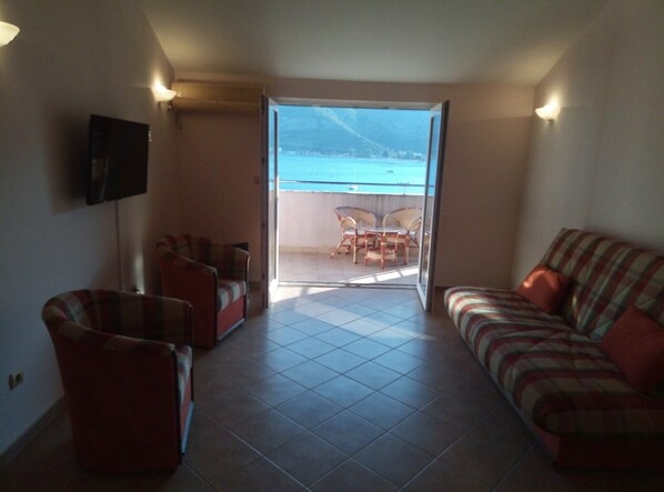 Una sala de estar con un sofá, sillones y un televisor en la pared. Hay una puerta corrediza de vidrio que da a un balcón con vista al mar.