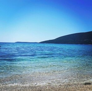 Una vista despejada de una playa con aguas poco profundas y piedras, una colina distante y un cielo claro.