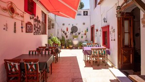 A narrow alley with tables and chairs under a red awning, surrounded by white walls adorned with various decorations and plants. - Ayvalık Bonjour Pansiyon (Ayvalik)