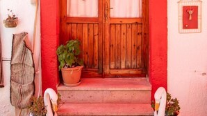 A red-framed wooden door with a number '7' above it, two potted plants on the steps, and a birdcage with a red bird on the wall to the right. - Ayvalık Bonjour Pansiyon (Ayvalik)