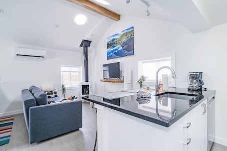 A modern kitchen with a black countertop, white cabinetry, and a built-in stove. There is a gray sofa with a colorful striped rug in the living area.