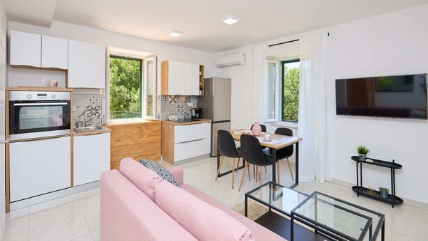 A modern kitchen with white cabinets, a dining area with a table and chairs, a pink sofa, and a TV mounted on the wall. - Apartment Fragola (Dubrovnik)