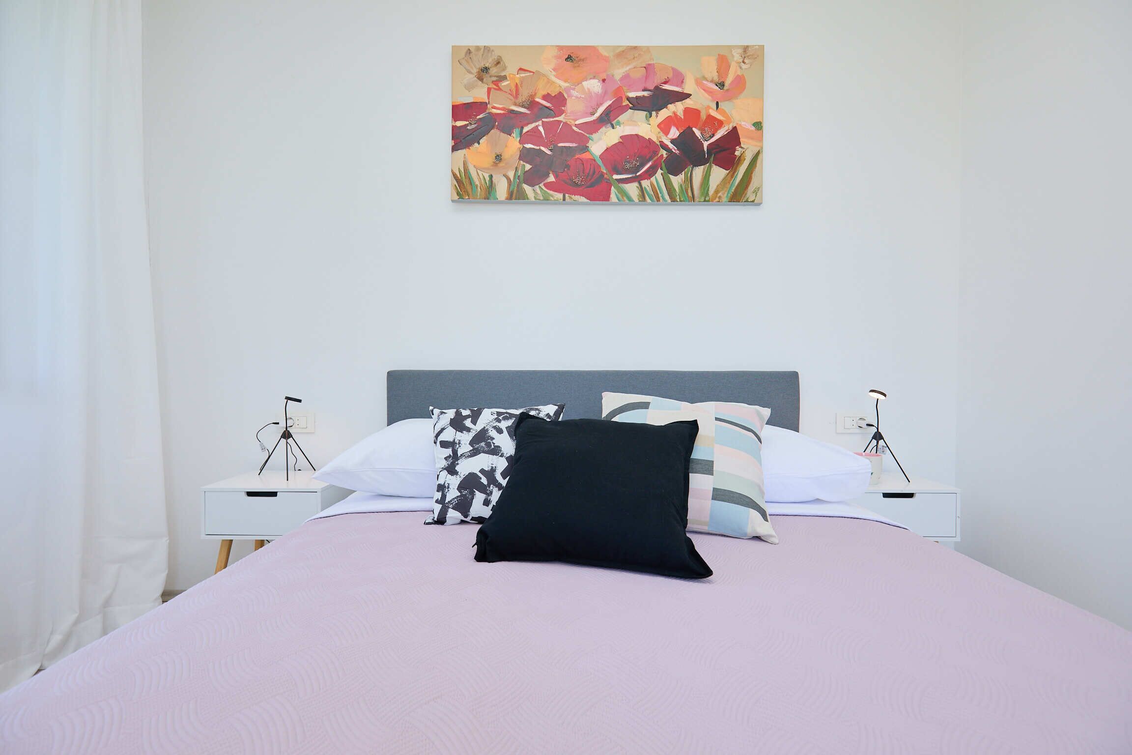 Een net opgemaakt bed met een roze dekbed en meerdere kussens, waaronder een zwart exemplaar, met aan weerszijden witte nachtkastjes en lampen.