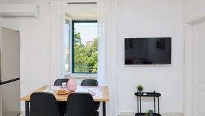 A modern kitchen with a built-in oven, a dining table with chairs, a TV mounted on the wall, and a window with curtains. - Apartment Fragola (Dubrovnik)
