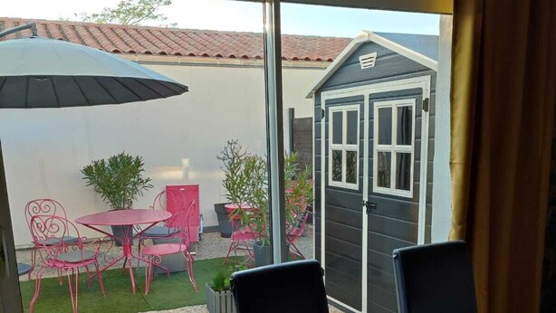 A room with a glass door leading to a patio area. There is a table with chairs, a small garden, and a shed visible through the door.