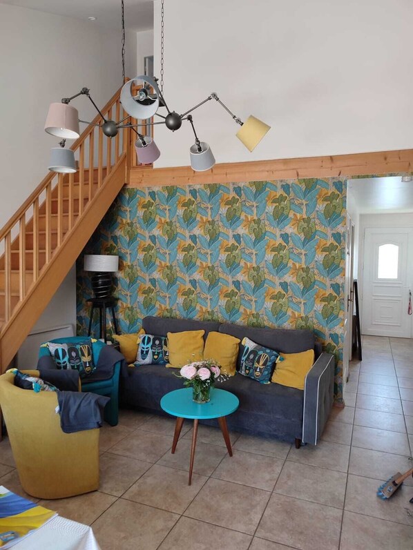 A living room with a staircase, a sofa with colorful cushions, a round coffee table, and a patterned wall.