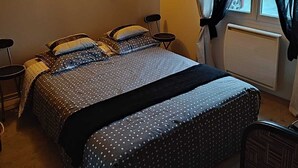 A bedroom with a bed, two chairs, a nightstand, a wall-mounted lamp, and a window with curtains.