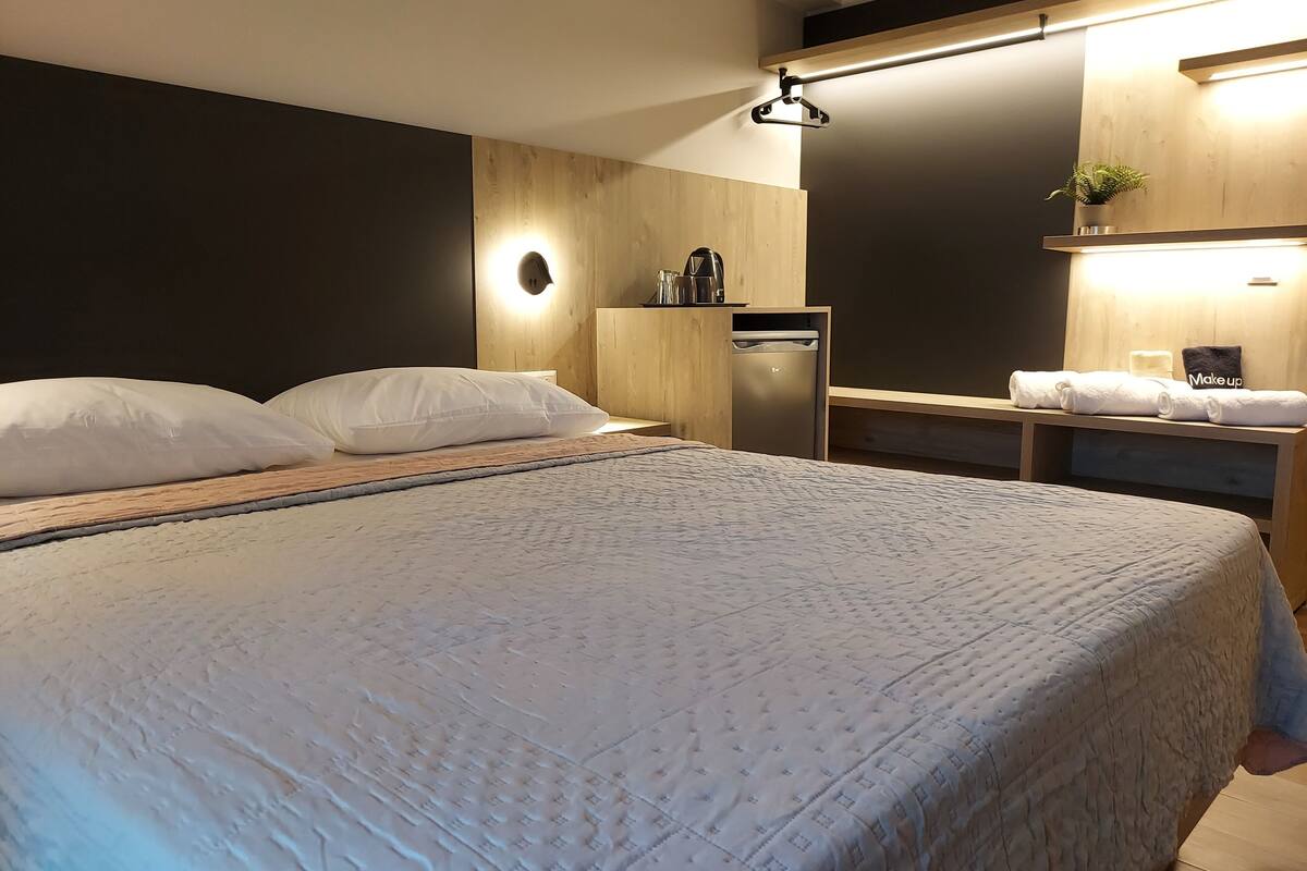 A neatly made bed with a quilted cover, a bedside table with a kettle and cups, a small shelf with towels, and a wall-mounted light fixture.
