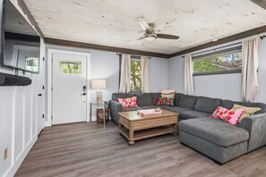 Un salon avec un canapé d’angle gris, une table basse en bois, un ventilateur de plafond et une porte avec une fenêtre.