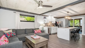 Una sala de estar moderna con un sofá gris, una mesa de centro de madera y una cocina con armarios blancos y un taburete de bar.