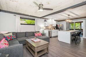 Ein modernes Wohnzimmer mit einem grauen Sofa, einem Holztisch und einer Küchenzeile mit weißen Schränken und einem Barhocker.