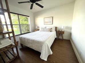 A bedroom with a bed, a wooden nightstand, a lamp, a ceiling fan, and a window with blinds.