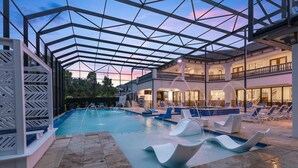 Un área de piscina con techo de cristal, sillones de descanso y un edificio con varios balcones.