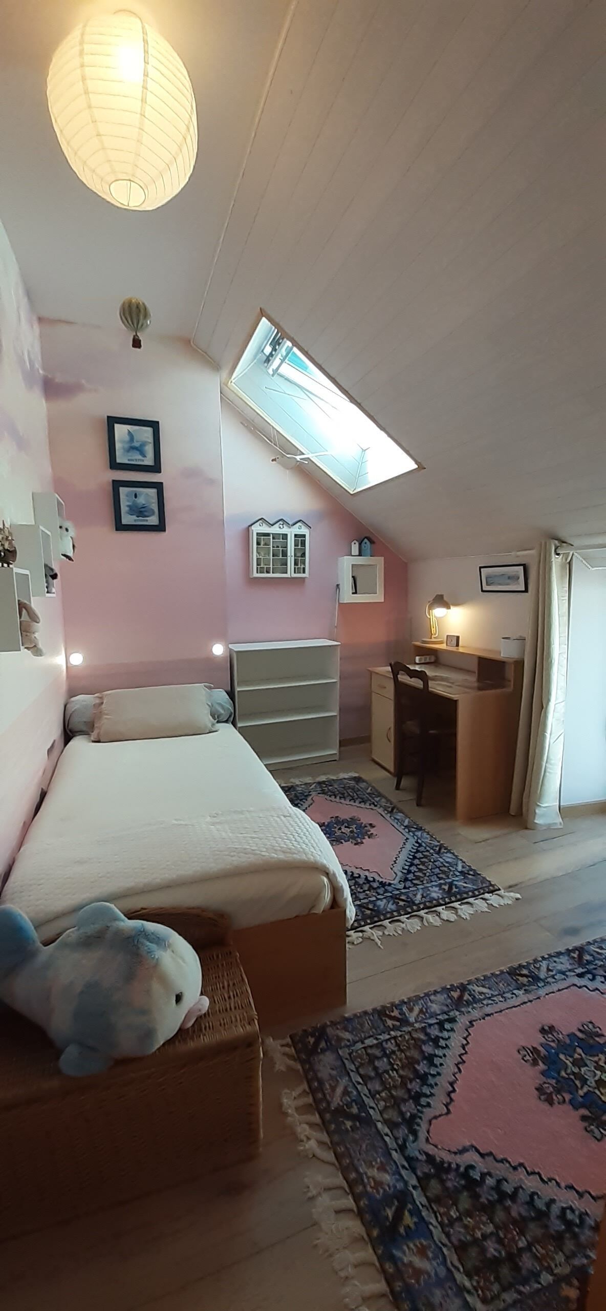 A bedroom with a bed, a desk, a chair, a small table, a lamp, a window, and a ceiling light.