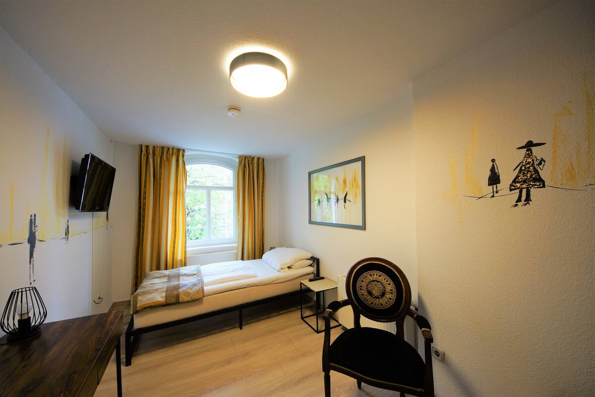 A bedroom with a single bed, a chair, a desk, a television, and a window with curtains.