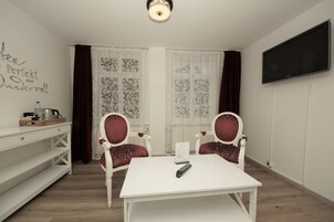 Quarto com mesa branca, duas cadeiras estampadas, prateleira branca, TV de tela plana e janela com cortinas.