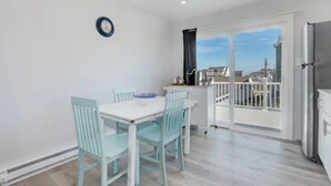 A dining area with a white table and light blue chairs, a kitchen with a refrigerator, and a balcony with a view of buildings.