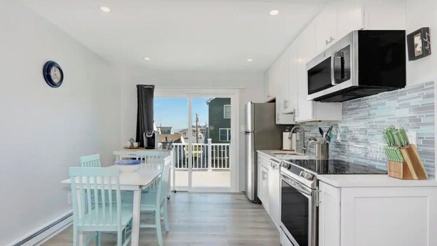 A modern kitchen with white cabinets, a black microwave, and a tiled backsplash. There is a dining area with a white table and light blue chairs.
