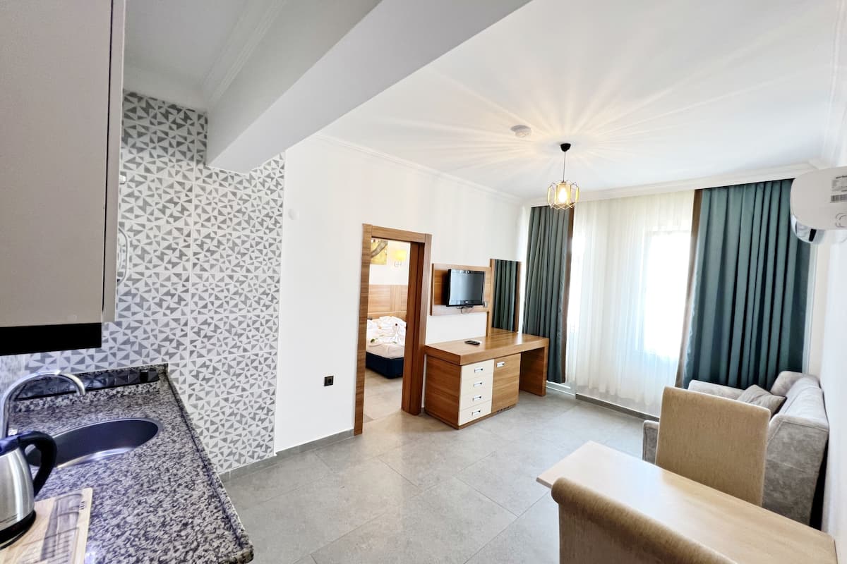 A modern hotel room with a kitchen area, a seating area with a sofa and armchair, a wooden desk, and a television.