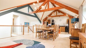 A spacious dining area with a high ceiling and exposed wooden beams, a kitchen in the background, and a colorful abstract rug on the floor. - Polurrian House (Helston)