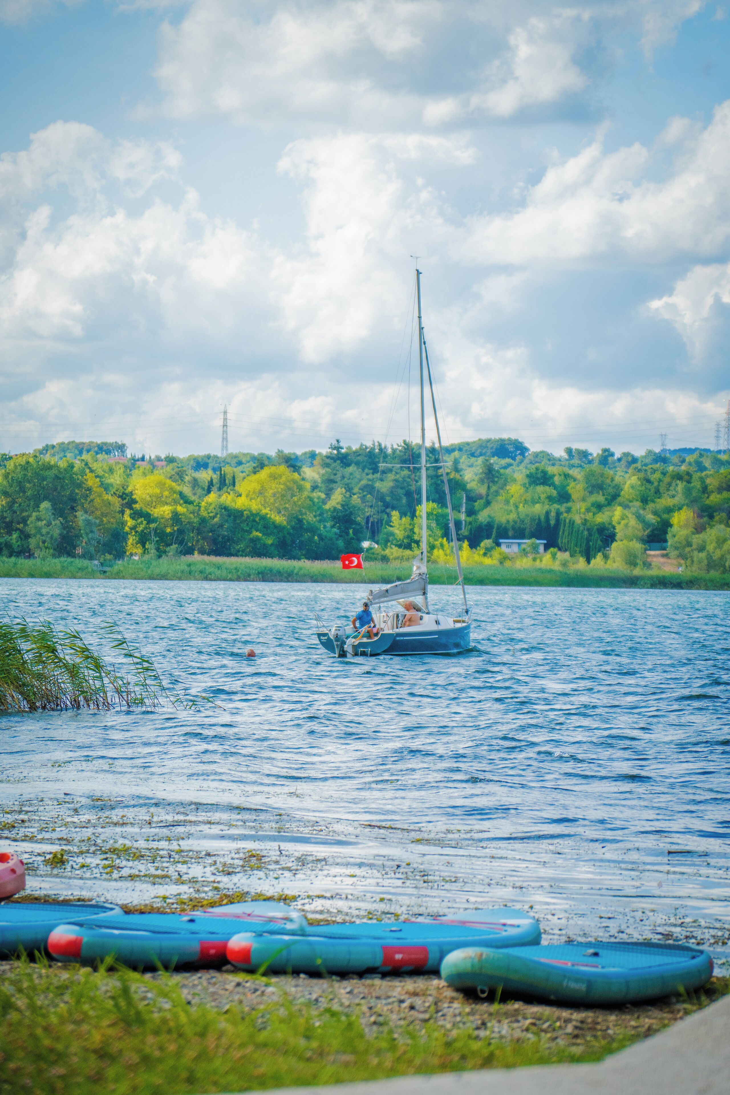 A calm lake with a sailboat, surrounded by greenery and a clear sky.