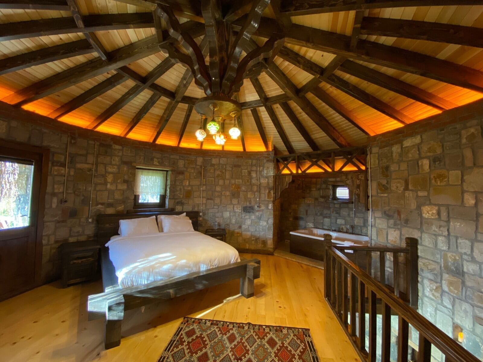 A rustic bedroom with a stone wall, a wooden ceiling, a bed, a nightstand, a small table, and a staircase.