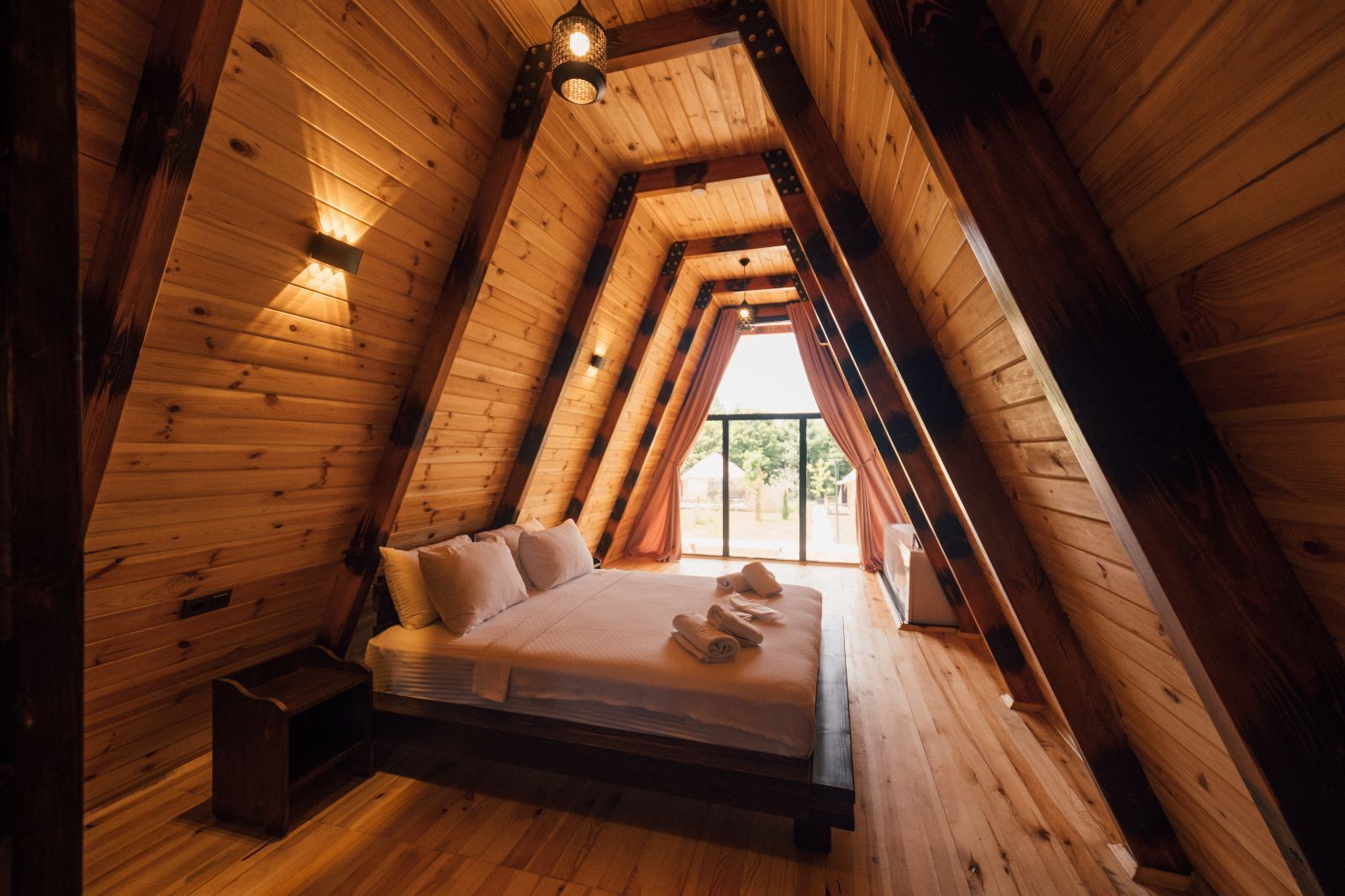 A cozy attic bedroom with a wooden sloped ceiling, a bed with pillows, and a small bedside table.