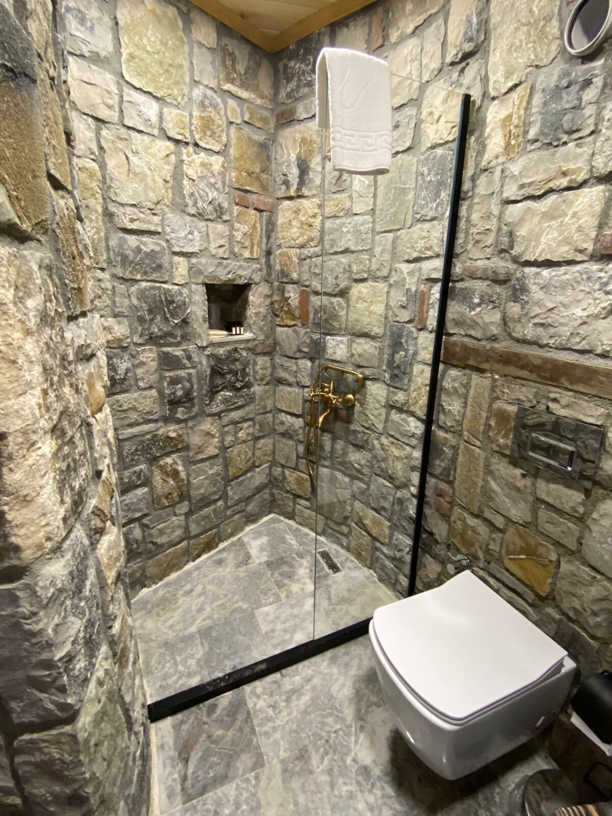 A stone-walled shower with a glass door, a white toilet, and a small shelf with toiletries.