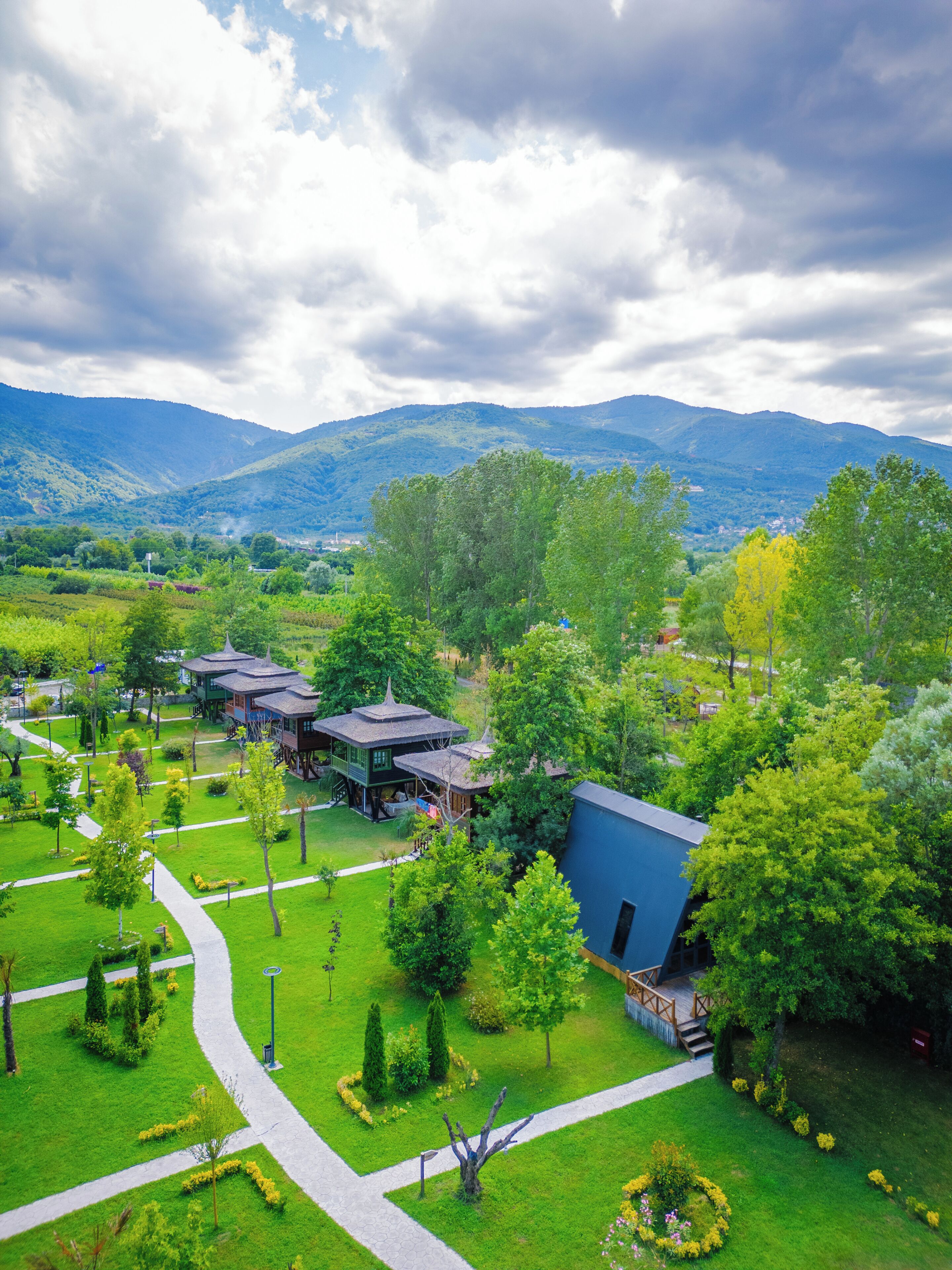 A park with pathways, a gazebo, and a blue building surrounded by greenery and mountains.
